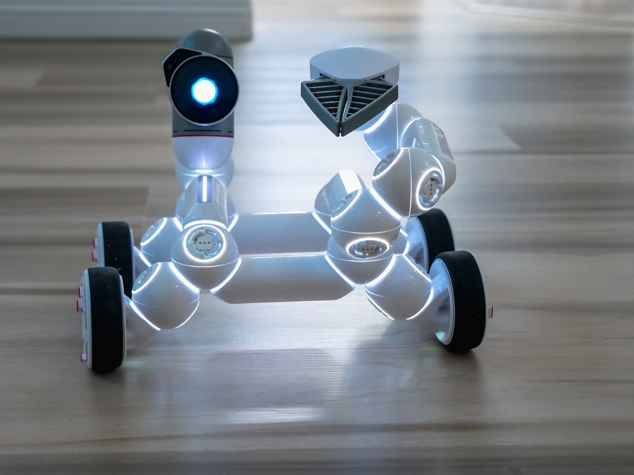 A close-up of a wheeled robot with illuminated joints and wheels on a wooden floor indoors.